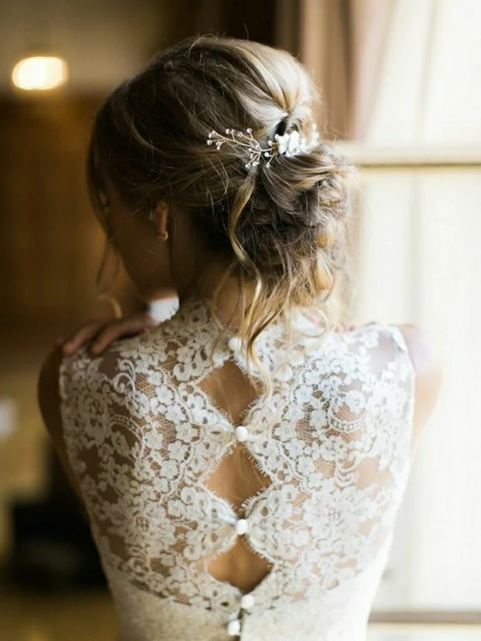 close up of a blonde bride facing backwards, wearing her hair in a messy bun with white ornaments, and a white dress with button up back made of sheer lace