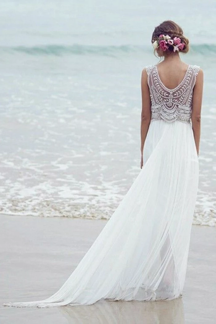 simple wedding dress, woman in long white dress with pleats and a lacy embroidered back, standing on a sandy shore in front of the sea, formal up-do with flowers in hair
