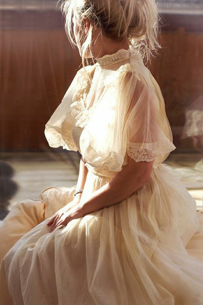 unique wedding dresses, blonde woman sitting down with hands in lap, in a Victorian cream wedding dress with lace and sheer short sleeves, messy hair bun
