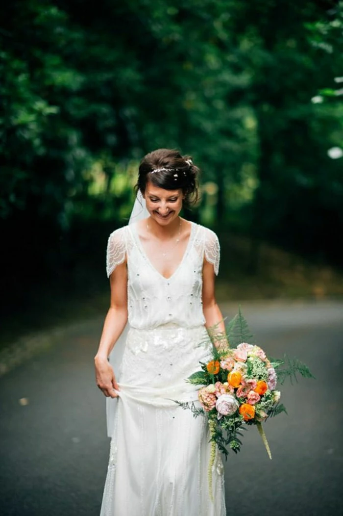 smiling woman in loosely fitting bridal gown inspired by 1920s fashion, looking down and holding her dress in one hand and a bouquet of colorful flowers in the other, trees and a road