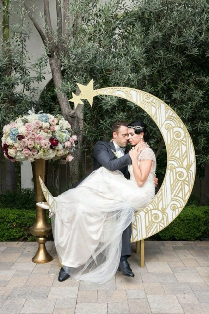 woman in flowing white bridal gown with lace and tulle, sitting on a moon-shaped ornament, hugged by a man in a dark grey suit, near a large vase with flowers, trees in background