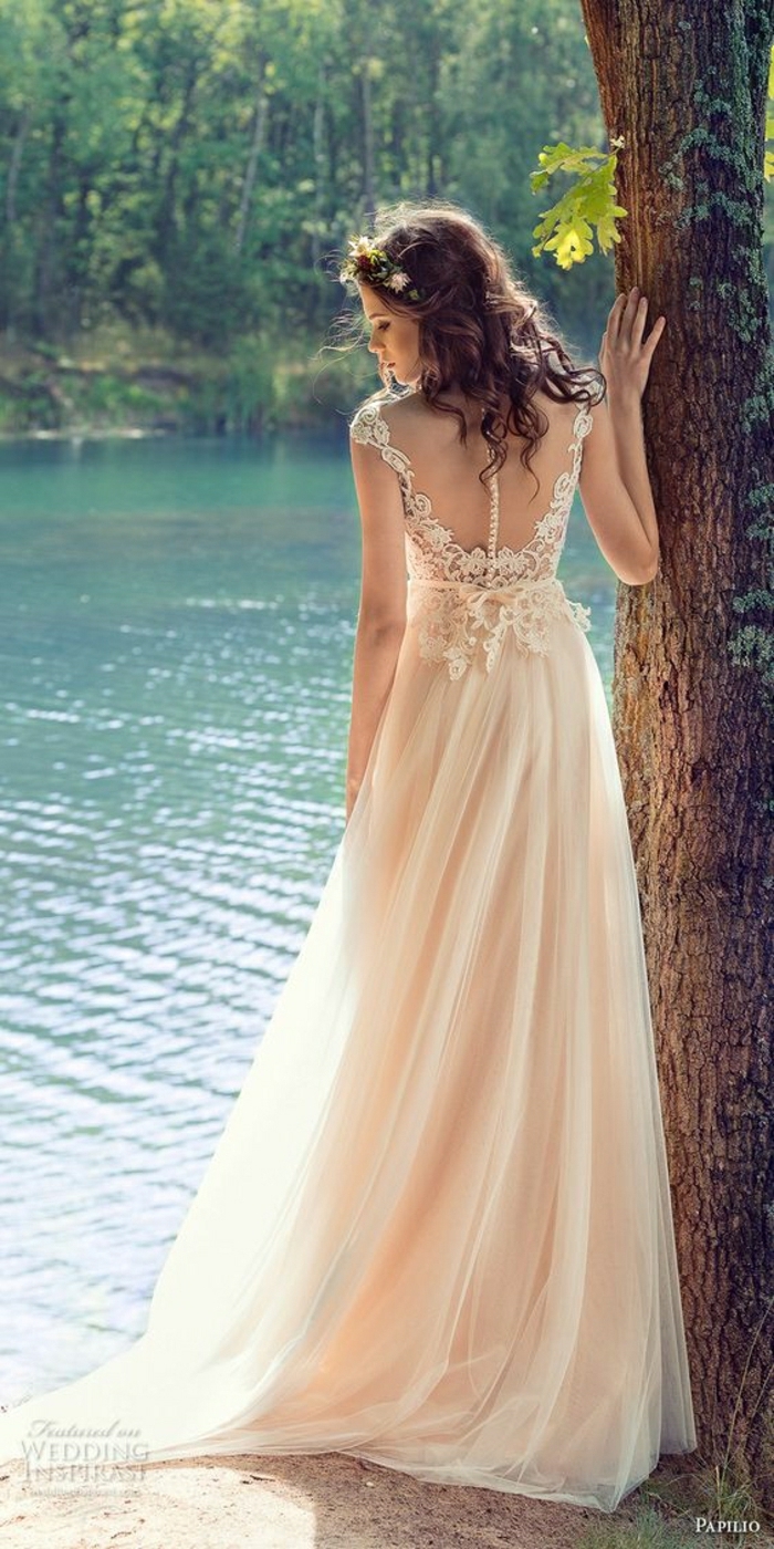 simple wedding dress, young bride with curly dark hair and a small flower crown, with a long cream wedding dress with lace details near the back, one hand on a tree and standing near a woodland lake