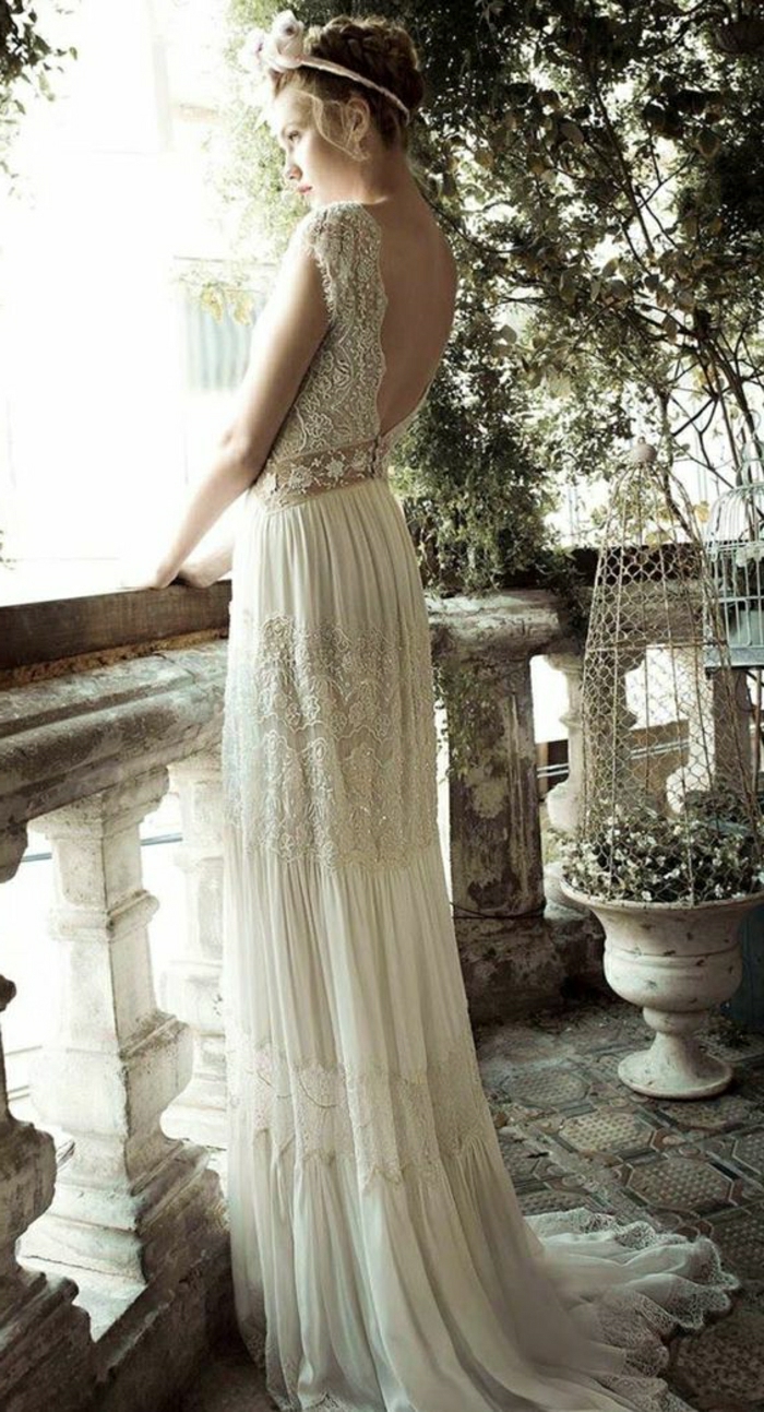 young blonde bride in vintage lace wedding gown with open back, with white hair ornament, standing on an antique looking balcony with trees and plants in the background