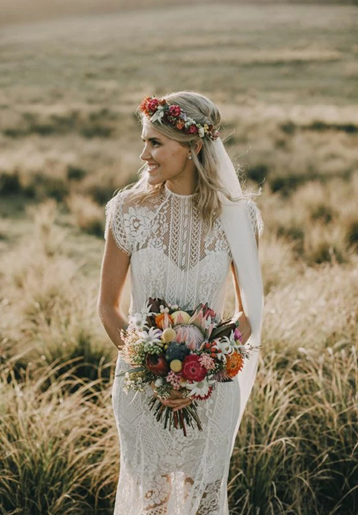 smiling bride with free-falling shoulder-lenght blonde hair, looking sideways, standing in a yellow field, dressed in a white long partly see-through dress with lace, holding a colorful bouquet and wearing a flower crown attached to a veil