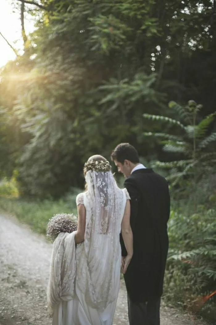 bride wearing long vintage lace dress, flower crown and veil and groom in black suit, both facing backwards, walking on a country road surrounded by trees,