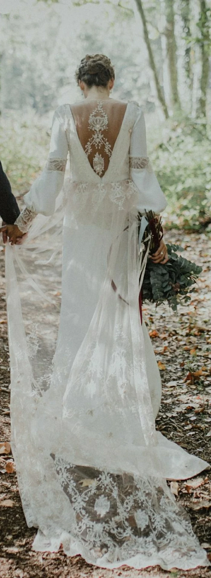 long white bohemian wedding dress with wide sleeves, cutout lave back and long veil, worn by a brunette with a fancy hairdo, holding a man's hand and a bouquet of flowers, woodland background
