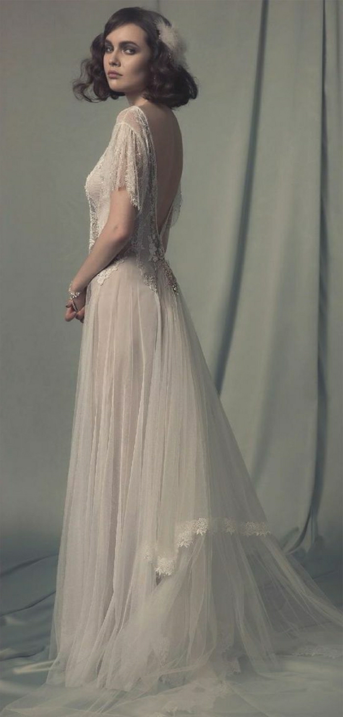 vintage inspired wedding dresses, young woman with 1920s style hair and a white hair ornament, wearing a long white embroidered wedding dress with low back and lace details