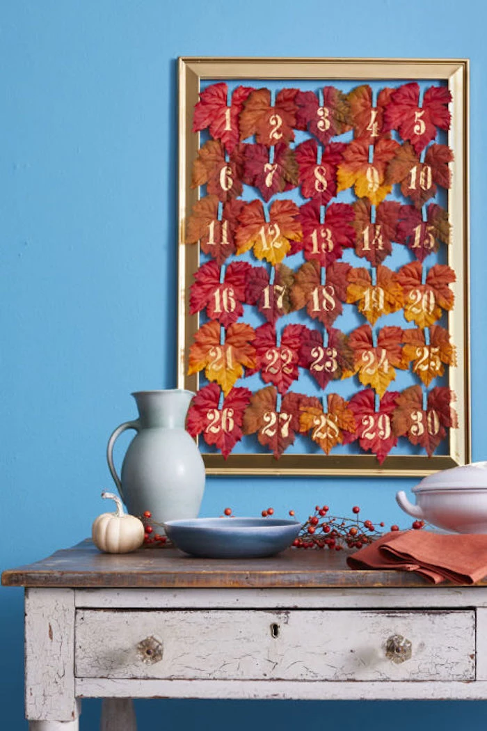 golden frame containing thirty numbered, colorful autumn leaves, hung on a blue wall, near a white and brown antique table with a drawer, blue pitcher and plate, brown dish, orange napkins, a small pumpkin and decorative berries on a twig