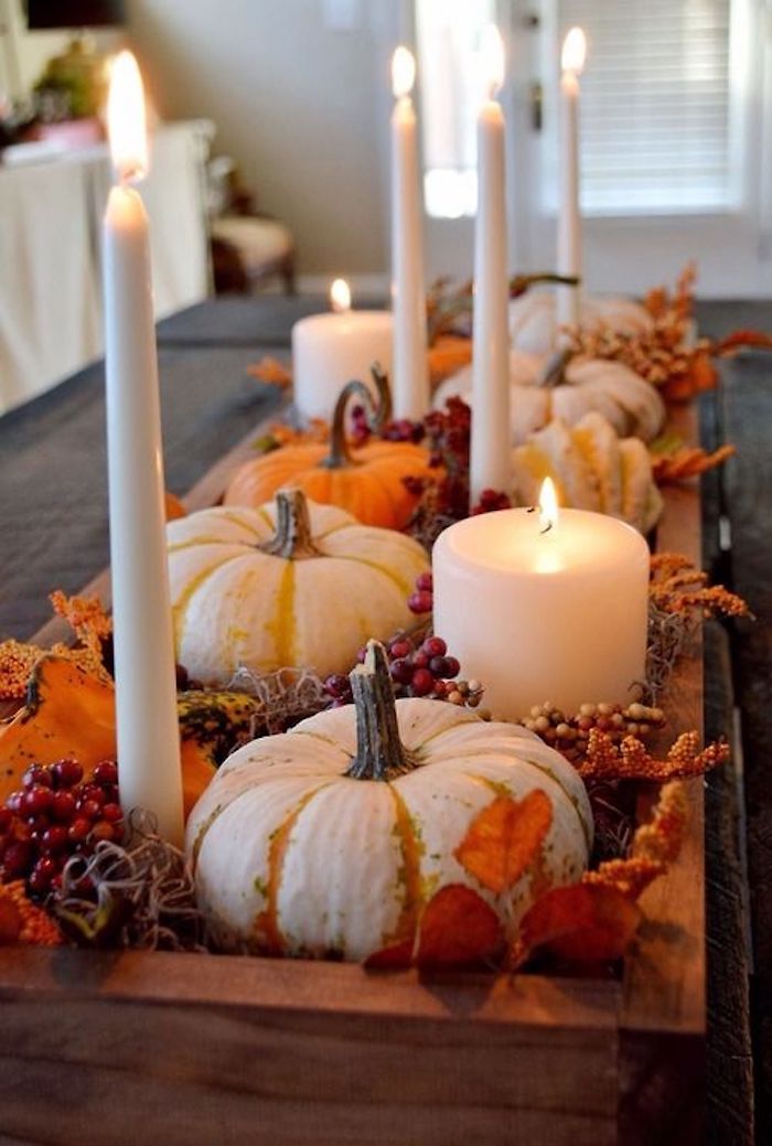 pumpkins in white and orange, placed on a wooden tray, with lit white candles, orange autumn leaves, decorative berries, on a dark table, room in the background