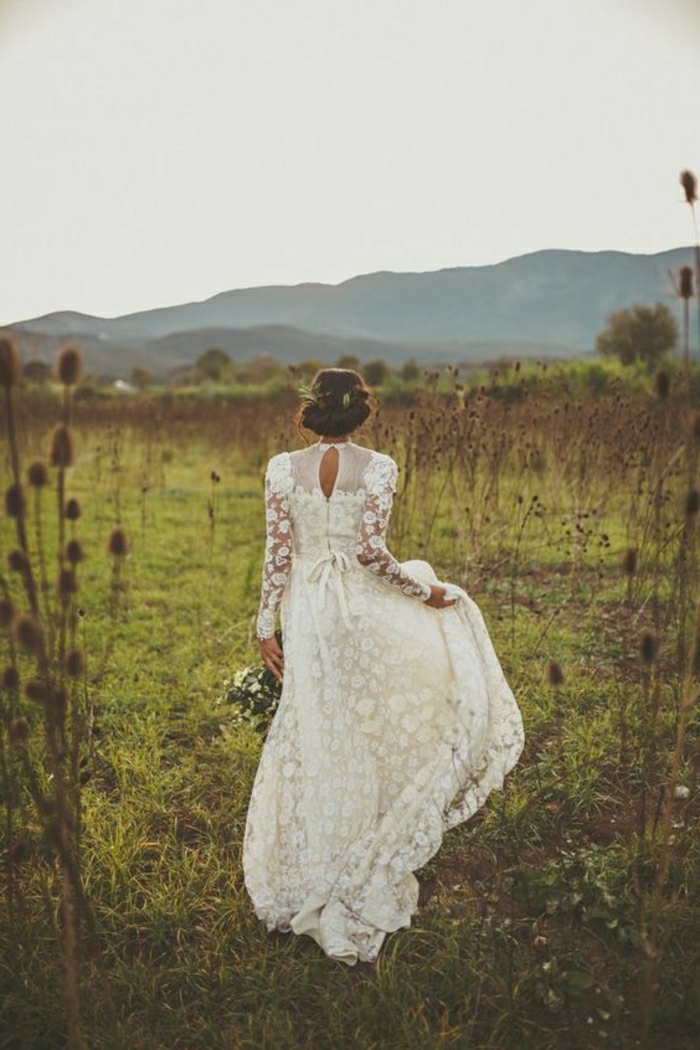 woman with brown hair in a fancy do, wearing a long vintage inspired dress with lace and sheer details, walking in a green field with thistles, holding her gown in one hand and a bouquet of white flowers in the other