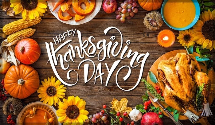 brown wooden table, happy thanksgiving day written in white fancy writing, plates of fruit and orange sauces, a cooked turkey, pumpkins, sunflowers, garlic, berries, corncobs, grapes, apples, chestnuts, pomegranate, orange and yellow autumn leaves