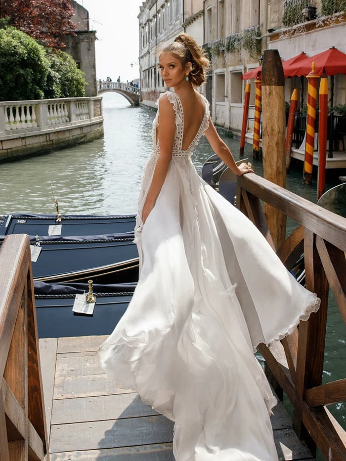 vintage wedding dresses, young bride with tied back hair, standing on quay in Venice near boats and a bridge, long white dress floating in wind, with lace details