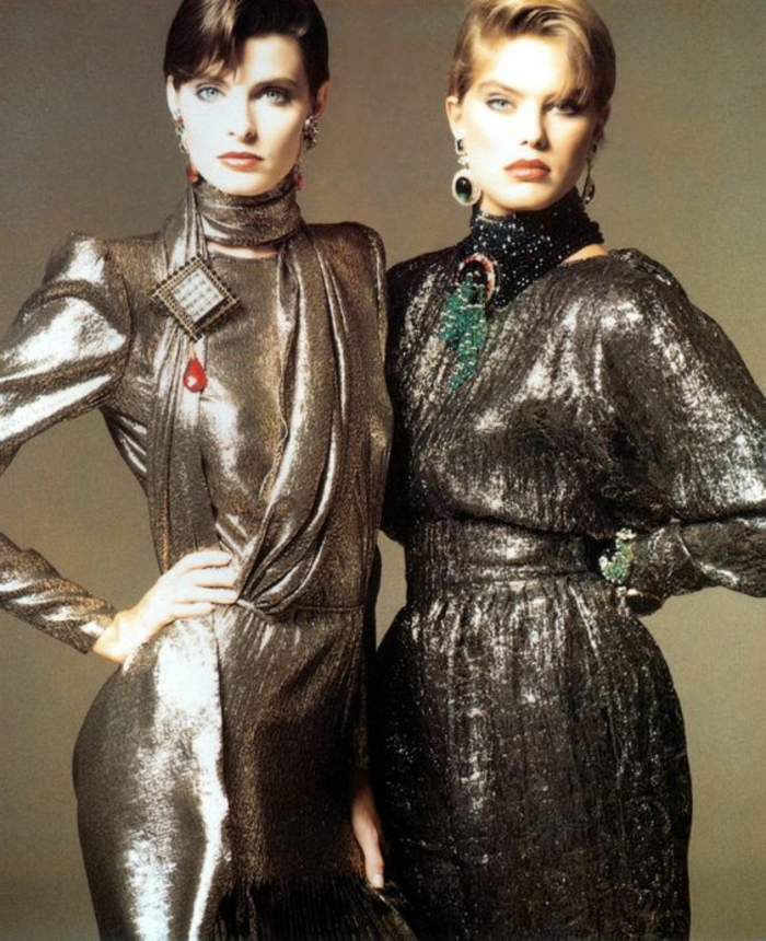 throwback outfits, two women with short brown and blonde hair standing next to each other, hands on hips and wearing 80s shiny silvery dresses with scarves and jewelry