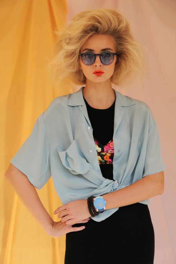 80s clothing, young woman with feathery died blonde hair, light blue short-sleeved tied shirt, black dress with flower embroidery, hands on hips and yellow curtains