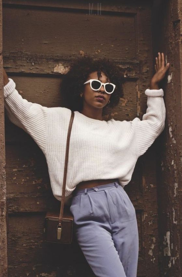 80s fashion, african-american girl leaning on old door, with black curly hair and brown bag, white ribbed sweater with rolled-up sleeves, sunglasses with white plastic frames, pale lilac carrot pants