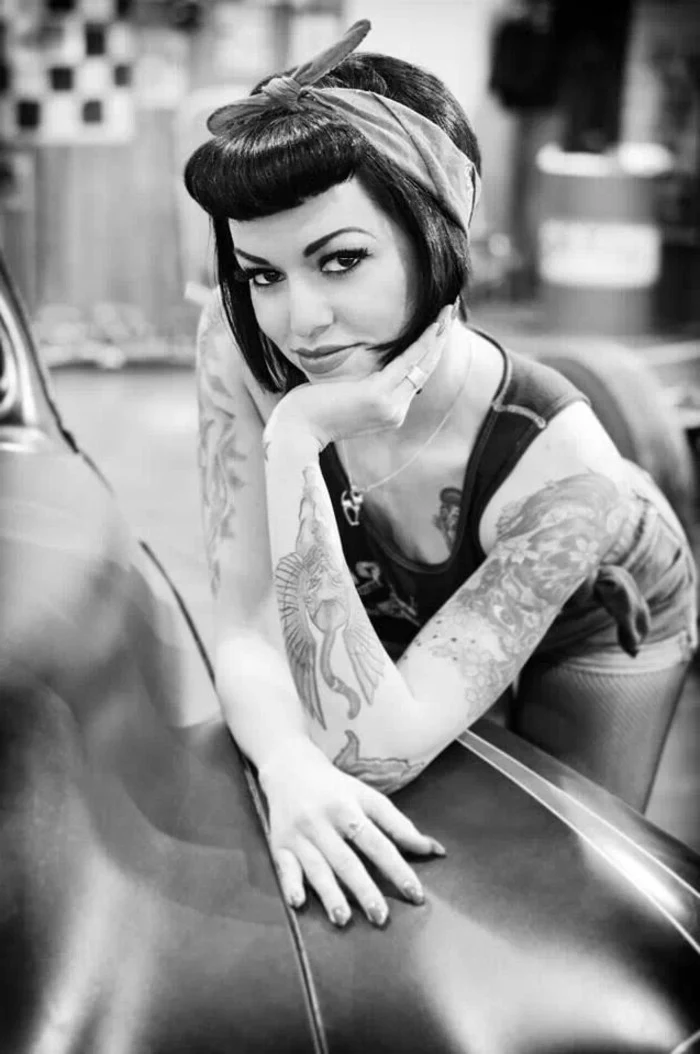 pinup hair, woman with short black hair tied with bandanna and rolled bangs, leaning on vintage car, many tattoos on arms and body