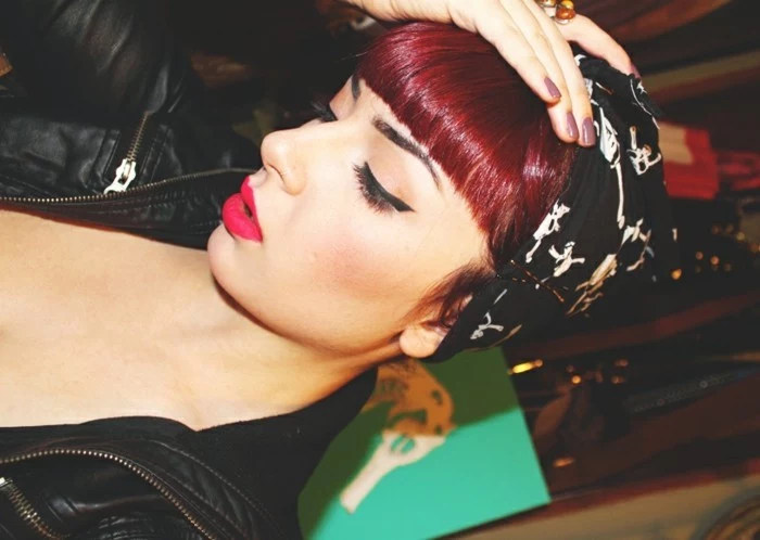 bettie bangs, sideways photo of woman with burgundy hair and bangs, black eyeliner and dark hot pink lipstick, wearing black leather jacket and black bandanna with white skull pattern