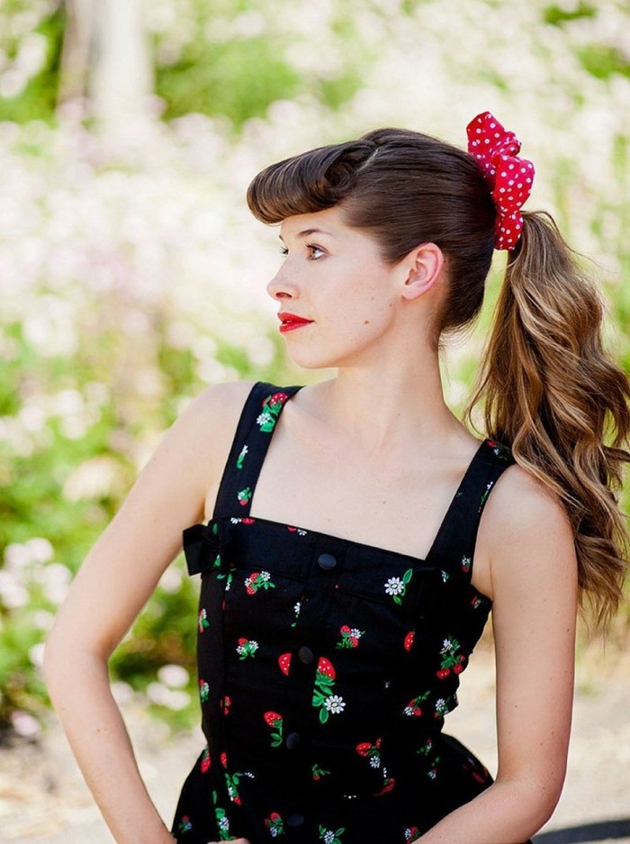pin up hairstyles, brunette woman in profile with rolled up bangs and a wavy ponytail, wearing a black sleeveless dress with strawberry pattern, red hair bow with white polka dots