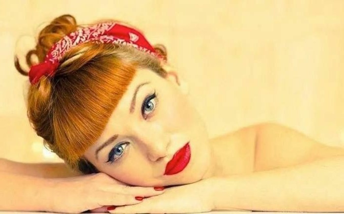 bettie bangs, ginger-haired woman with blue eyes and bold black eyeliner, vivid red lipstick and nail polish, red bandanna with white and black details, leaning on hands