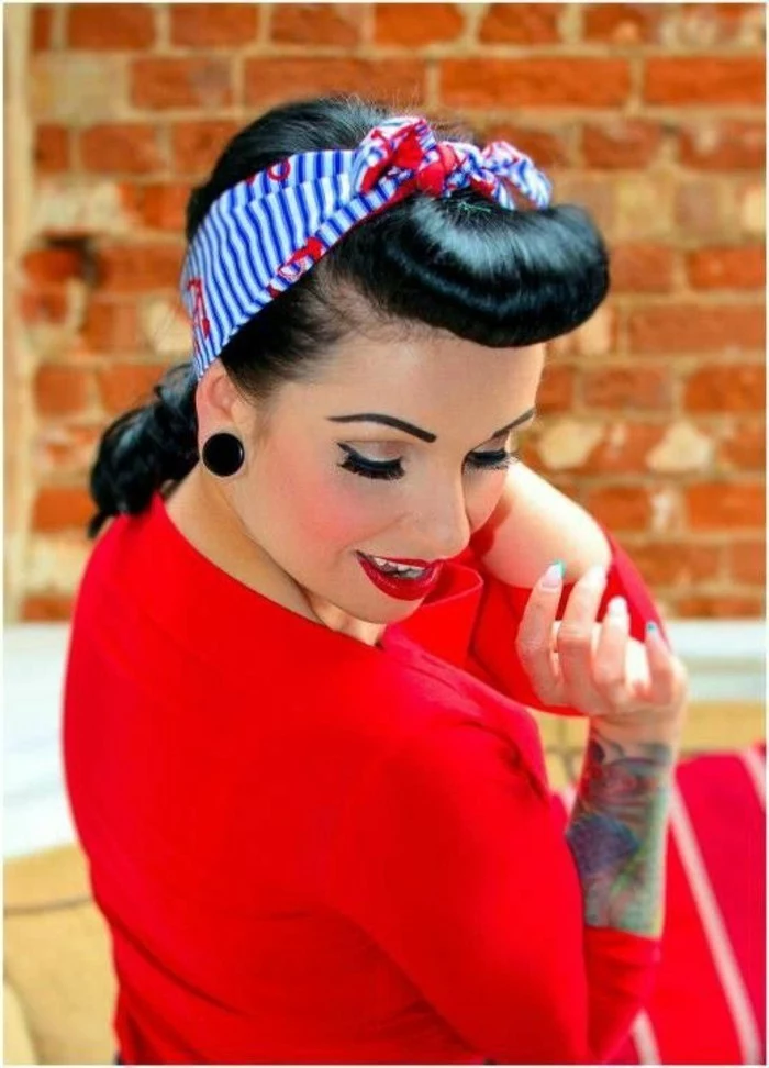 betty bangs, smiling black-haired woman seen from above, blue and white striped bandanna with red flowers, curled bangs, wearing red top and lipstick, fake eyelashes and mascara, big black earrings and tattoos on arm