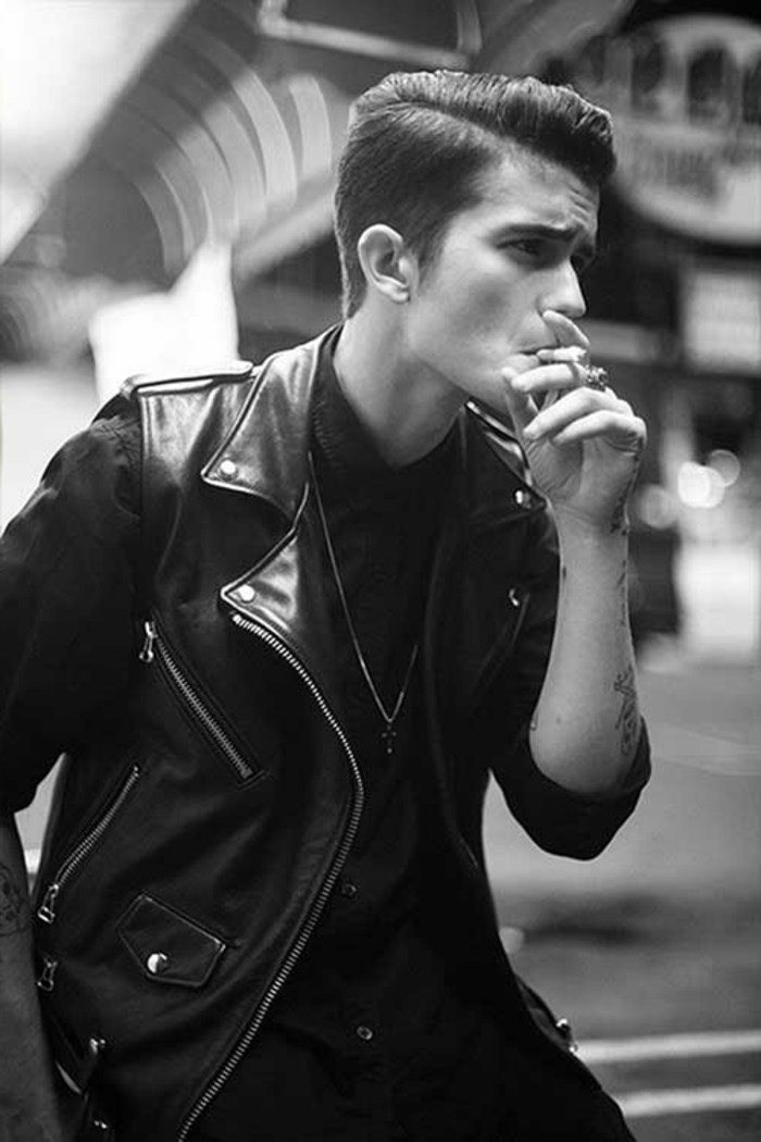 young man with gelled up parted hair, holding cigarette to his mouth, black leather jacket and tattoos, dark top and pendant