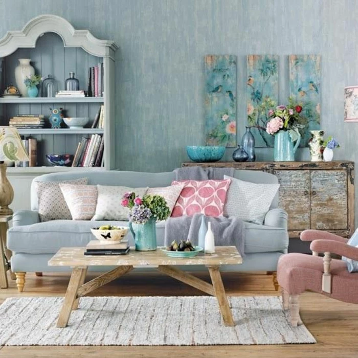 interior painting, pale blue wallpaper, pale blue pastel colored cupboard and sofa, six pastel colored cushions, pastel pink chair, pale wooden table and wooden floor, pale cream rug, artwork decorations and flowers