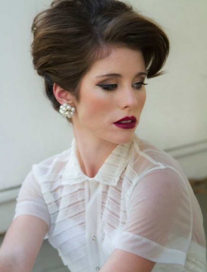 woman with dark red lipstick and heavy eye makeup, fake lashes eyeliner mascara, sheer white top and white cardigan underneath, pearl earrings and brown hair in retro updo