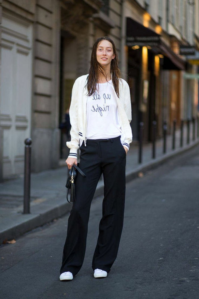 smiling brunette woman, wearing white baseball jacket, over white t-shirt with black print, with long black trousers and white sneakers, holding small black handbag