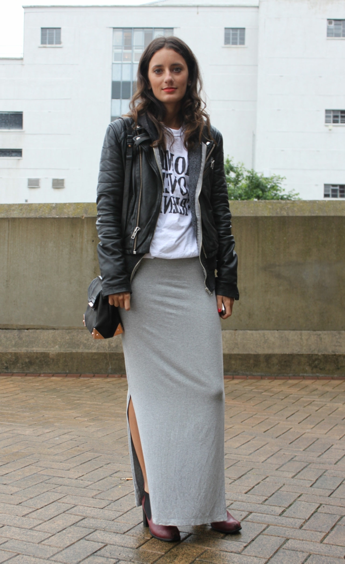 woman with curled brown hair and red lipstick, wearing big leather jacket, over white shirt with black print, grey jersey maxi skirt and ankle boots