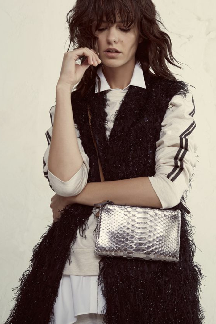 woman with messy brown hair and bangs, wearing long black fluffy fur black gilet, over white shirt with two black stripes on sleeves, small shiny silver snake leather bag