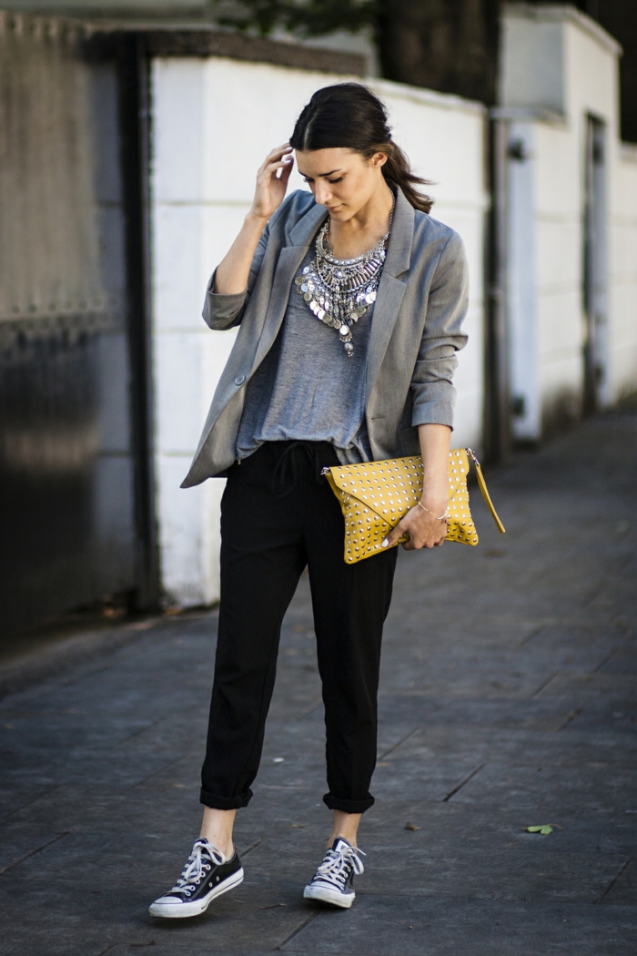 business casual attire for women, black ankle-length sports pants, grey top and grey blazer, plain sneakers and yellow clutch, worn by woman with dark hair and big elaborate silver necklace