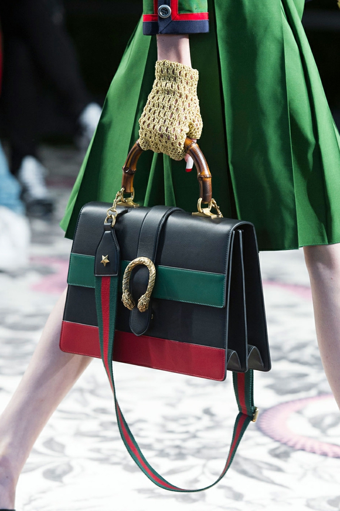 close up of black leather bag, green and red stripes, golden details and wooden handle, held by woman with yellow gloves, wearing green dress with red and blue details