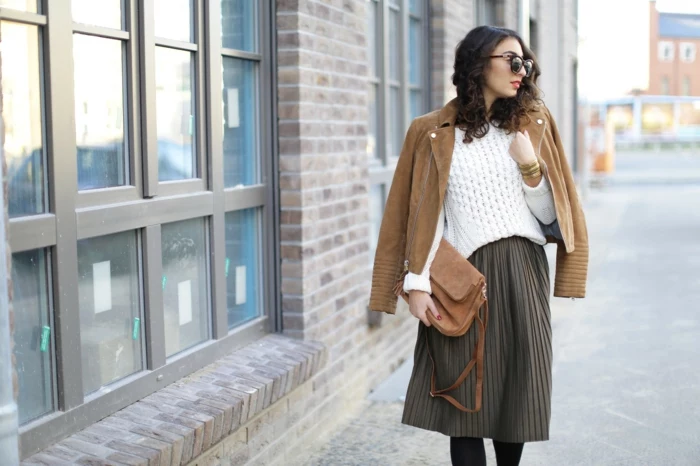 business casual attire for women, pleated knee-length brown skirt, white chunky knit sweater, camel brown leather biker jacket over shoulders, brown bag and sunglasses 