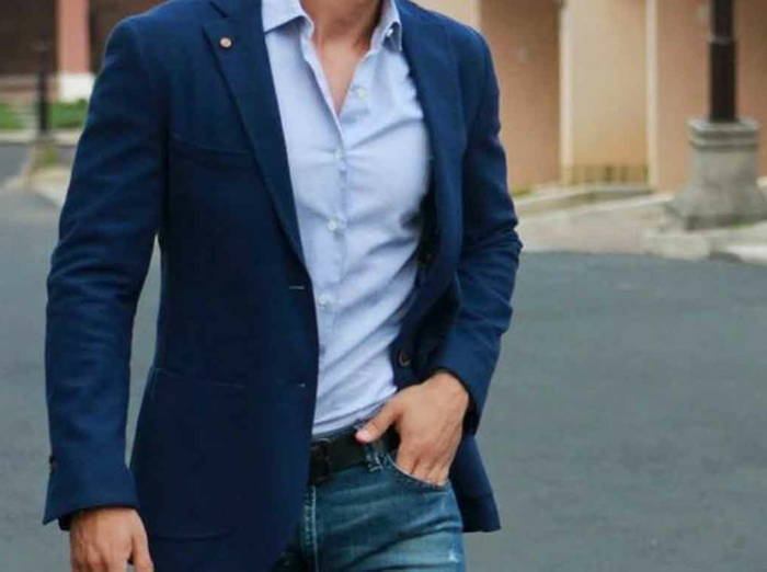 close up of male torso, dressed in dark blue blazer over light blue shirt, jeans with black belt and hand in pocket