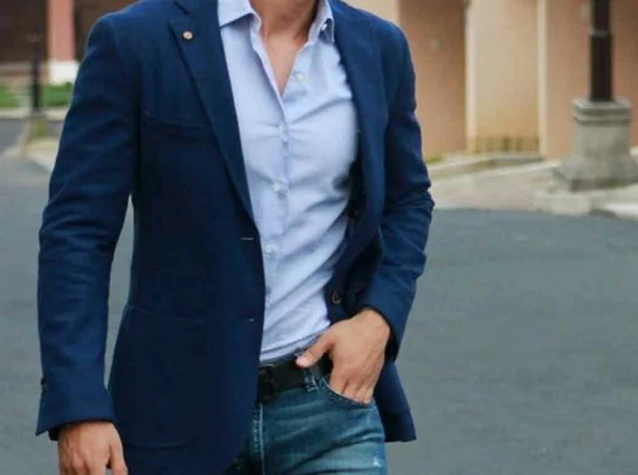 close up of male torso, dressed in dark blue blazer over light blue shirt, jeans with black belt and hand in pocket