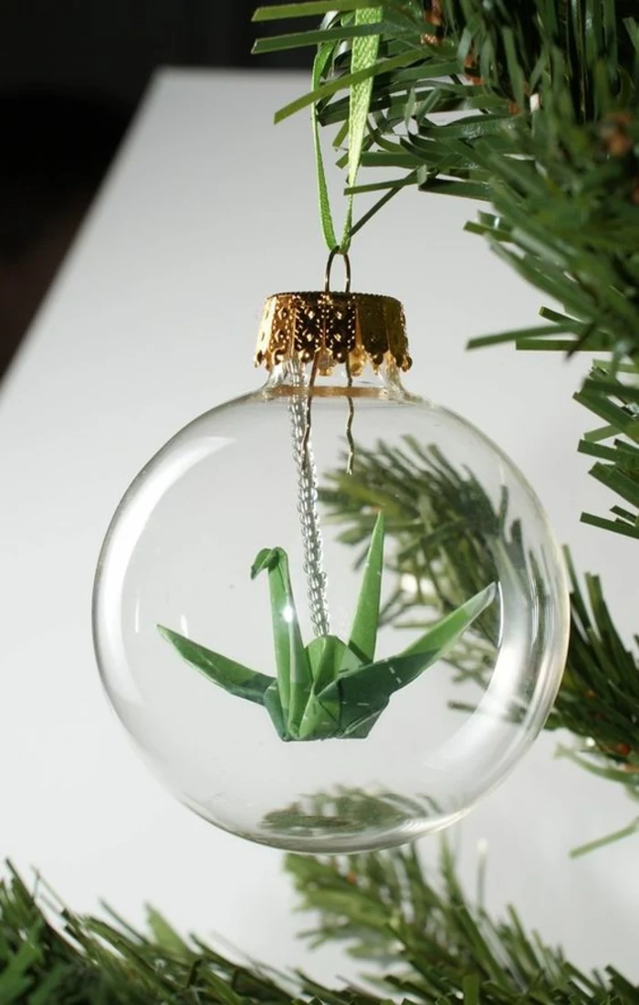 a small green paper origami crane, hanging on a small chain inside a clear glass bauble, golden cap attached to pine tree with green ribbon