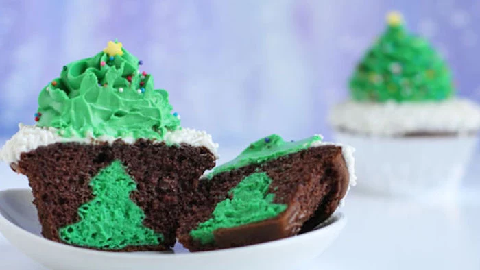 chocolate cupcake with white and green icing shaped like x-mas tree, cut in half to show green x-mas shape inside, on white plate with another cupcake in background