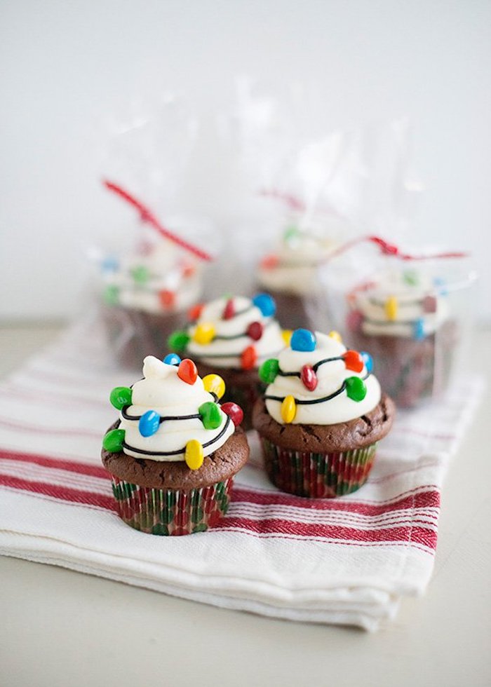 three chocolate cupcakes, with white frosting, decorated with chocolate drizzle and colorful candies made to look like christmas lights, three more wrapped cupcakes in background 