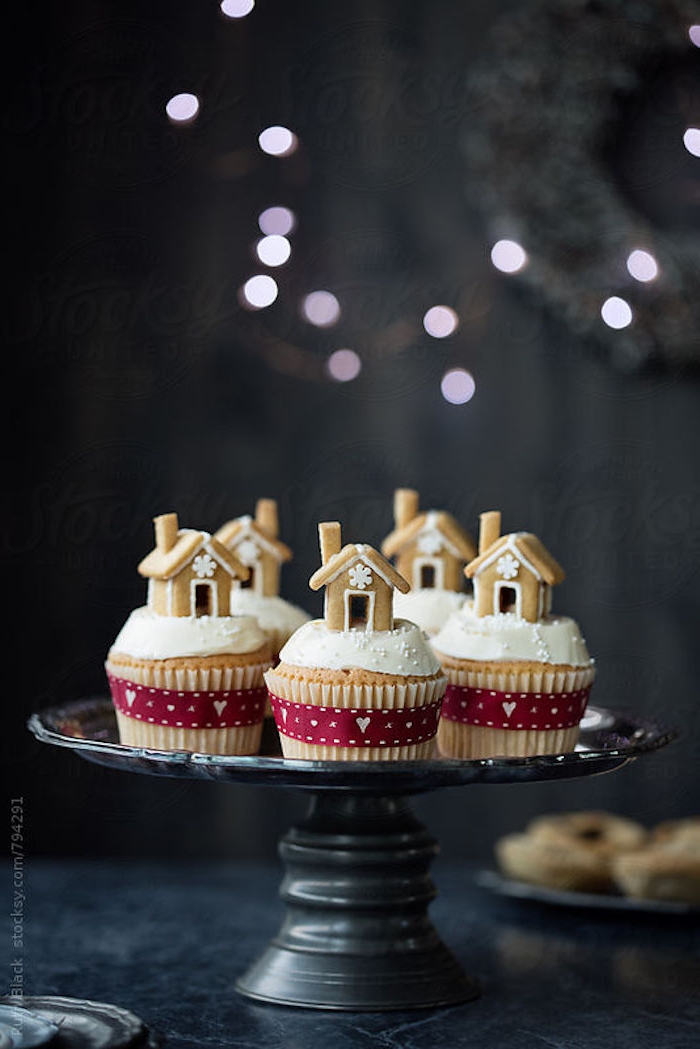 batch of cupcakes with white frosting, decorated with small gingerbread houses, tied with red festive ribbons, on black dish placed on table