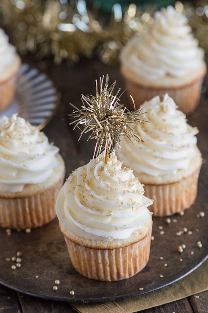 mini cupcake recipes, several light-colored cupcakes with white frosting, decorated with gold leaf pieces and golden-colored sparkler, on a dark plate with golden pearls