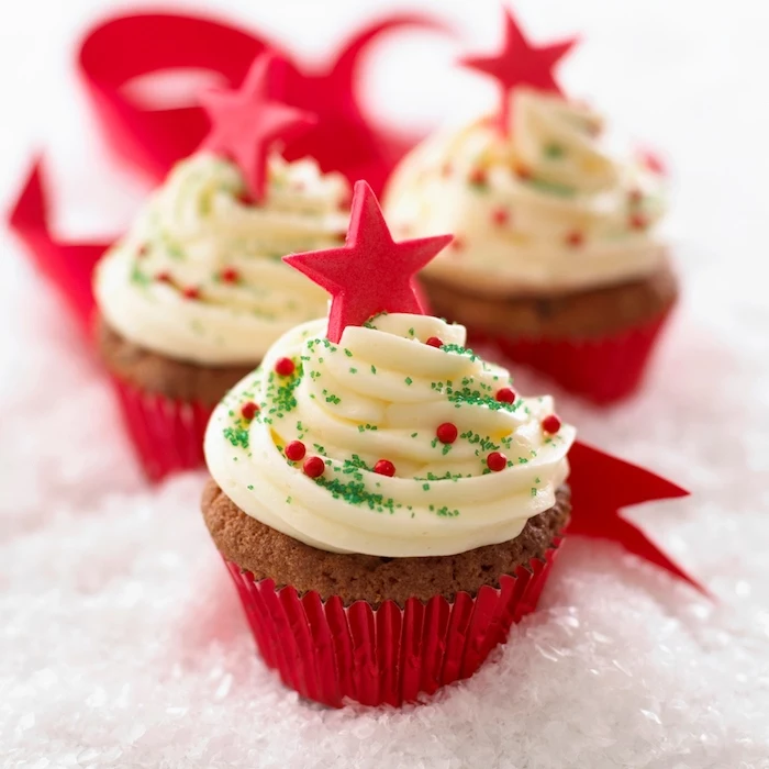 christmas baking ideas, three light brown cupcakes, pale yellow creamy frosting and red wrappers, decorated with red pearls and stars and green sprinkles