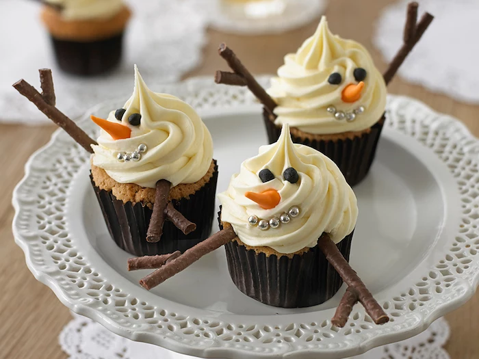christmas flavors, three cupcakes with dark wrappers, decorated with white frosting, with silver pearls, small fondant shapes and chocolate details, made to look like snowmen, on white plate