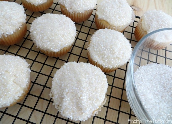mini cupcake recipes, several cupcakes with white frosting, decorated with sparkly white sugar sprinkles, near a glass bowl with more sprinkles, on a black metal oven grate