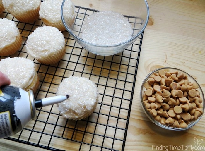 mini cupcake recipes, a hand holding can of spray chocolate icing to a cupcake, with white frosting and sparkly sprinkles, near bowls with sprinkles and caramel chips