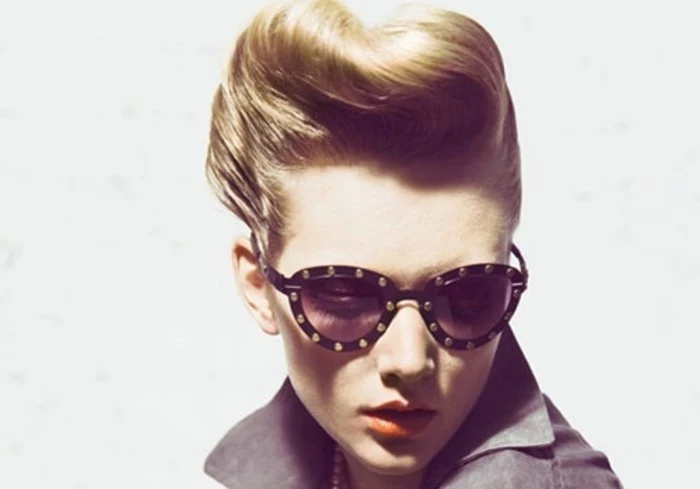 blonde woman with gelled up retro hair, dark sunglasses with metal studs, orange lipstick and a grey top, white background