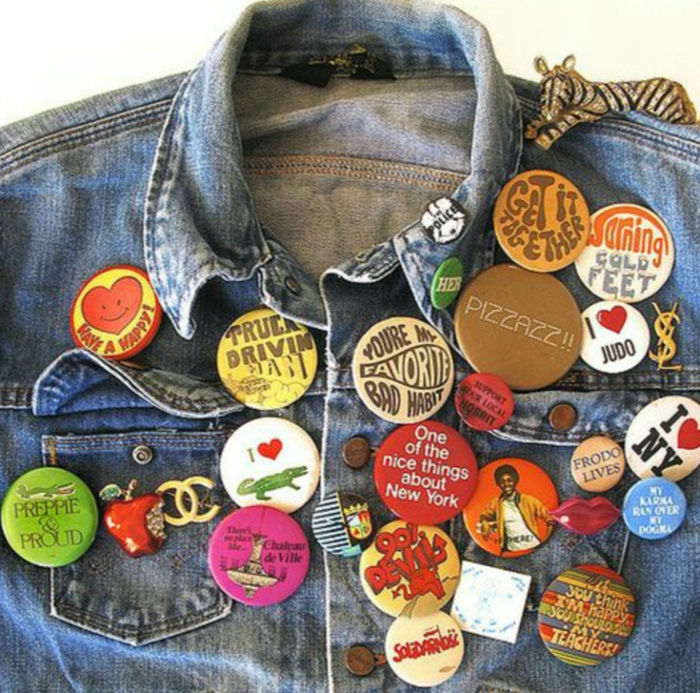 close up of a denim jacket with many retro pins, Chanel NY bitten apple and zebra ornament, white background