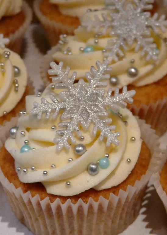 christmas flavors, close up of batch of vanilla cupcakes, pale yellow frosting, decorated with silver sparkly snowflake shape, pale blue and silver pearls