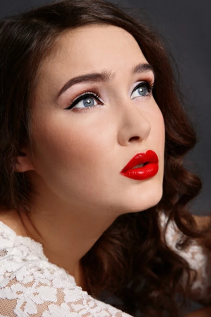 brunette woman with bold red lipstick, black eyeliner and fake lashes, curly hair and blue eyes, white lacy top