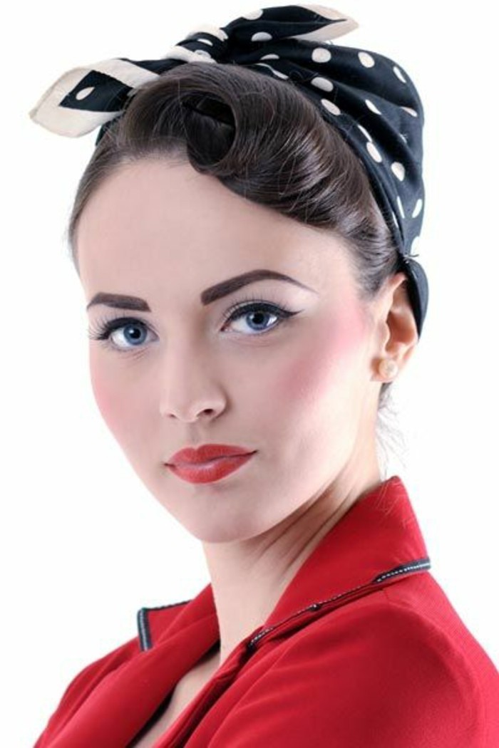 dark-haired woman wearing black bandanna with white polka dots, eyeliner mascara and blush, penciled eyebrows and red lipstick, red blazer with black details
