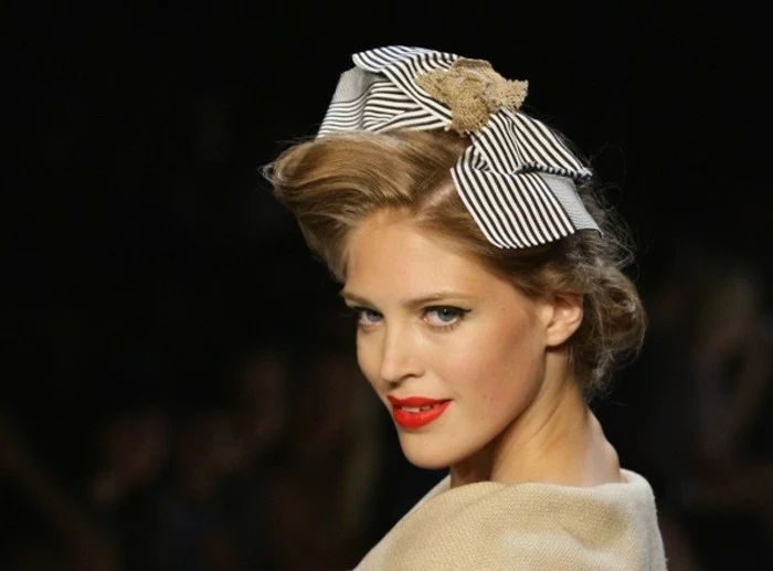 blonde woman close up, retro hairstyle with big striped hair bow, bright red lipstick and black mascara, camel-colored top and black background